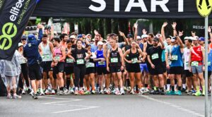 Group of runners at the starting line of a road race, bib numbers visible, hands raised in the air under a start banner.