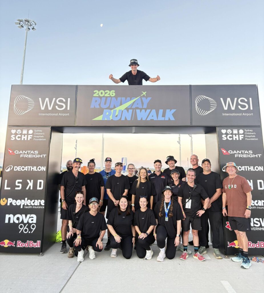 Group of people in black shirts posing on a stage with sponsor banners for a charity run/walk event, smiling at the camera. 2026 Runway Runwalk banner visible.