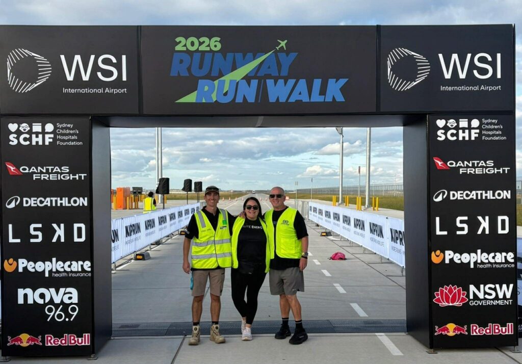 Three people in hi‑vis vests pose at a Runway Run/Walk 2026 finish arch at an airport, sponsor banners line the course behind them.