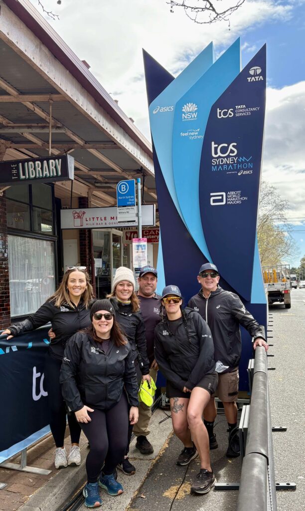 Group of runners in black jackets posing beside a tall blue TCS Sydney Marathon banner near shops.